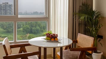 A dining area with a round table, four chairs, a fruit bowl, a potted plant, and a view of a cityscape through large windows.