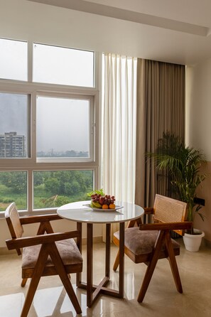 A dining area with a round table, four chairs, a fruit bowl, a potted plant, and a view of a cityscape through large windows. - Yashobhoomi Stay (New Delhi)