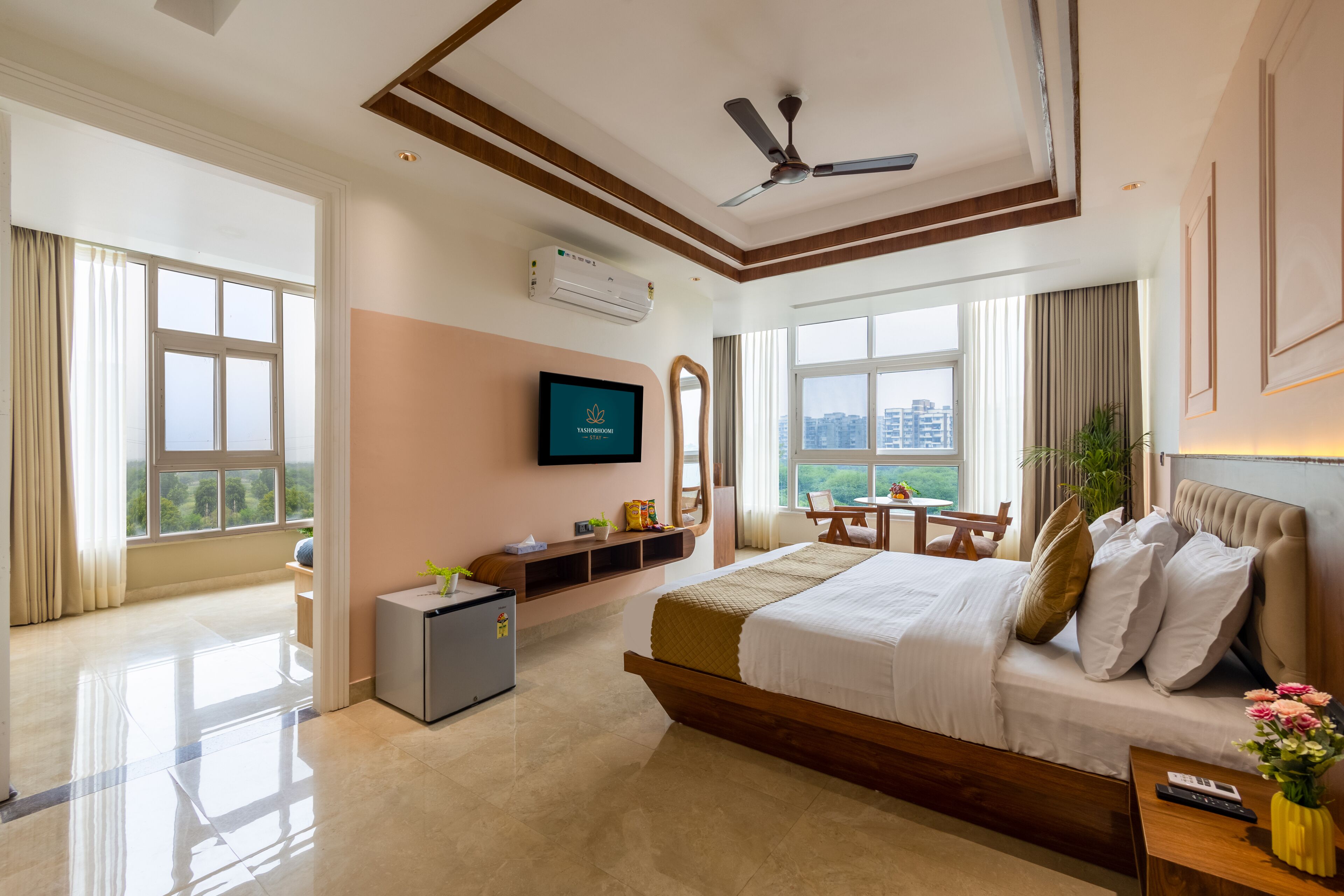 A hotel room with a bed, a television, a desk, a chair, a small table, a plant, and a view of the city.