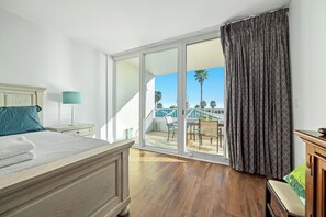 A hotel room with a bed, a nightstand, a lamp, a view of the outdoor area with a staircase and palm trees. - Caribe C213 - Gulf Front 3BR, Lazy River, Indoor/Outdoor Pools, 2 Min To Beach (Orange Beach)