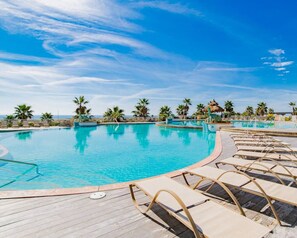 A large outdoor swimming pool with clear blue water, surrounded by lounge chairs and palm trees, under a bright blue sky. - Caribe C213 - Gulf Front 3BR, Lazy River, Indoor/Outdoor Pools, 2 Min To Beach (Orange Beach)