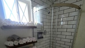 A bathroom with a glass shower enclosure, white tiled walls, and a window. There are shelves with folded towels and a white basket on the floor.