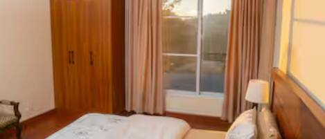 A bedroom with a large bed, a nightstand, a window with curtains, and a wooden wardrobe.