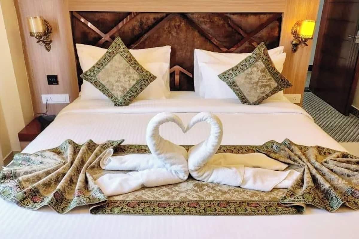 A hotel room with a large bed, two decorative pillows, a heart-shaped towel arrangement, and a patterned bedspread.