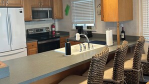 A modern kitchen with wooden cabinets, stainless steel appliances, a built-in island with bar stools, and a window with blinds. - Kraken's Cove: 4‑bedroom Ramrod Key escape, minutes from open water (Summerland Key)