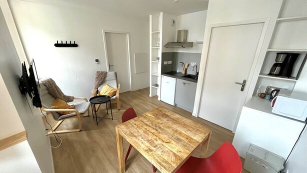 Cozinha compacta com mesa de madeira e cadeiras vermelhas, geladeira branca e pequeno espaço com pia.