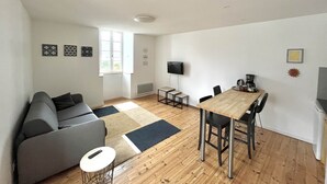 Een moderne woonkamer met een bank, salontafel, eettafel met stoelen, een aan de muur bevestigde televisie en een raam dat voor natuurlijk licht zorgt.