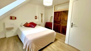 Een slaapkamer met een schuine kap, een bed met een witte spreidek en rode kussens, een houten garderobe en een witte deur.