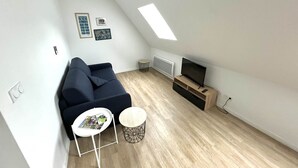 Een moderne loft-achtige woonruimte met een hoge plafond, een donkerblauwe bank, een klein rond tafeltje, een televisie op een houten dressoir, en een dakraam.