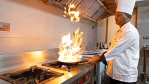 A chef performing a flambé with a pan on a stove in a kitchen. - Yashobhoomi Hostel  (New Delhi)