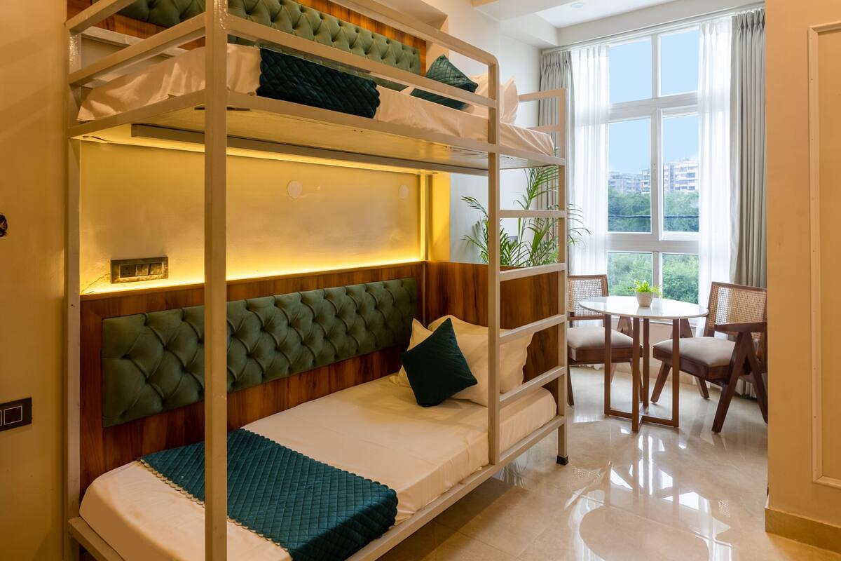 A bunk bed with a cushioned headboard, a small table with chairs, and a window with curtains.