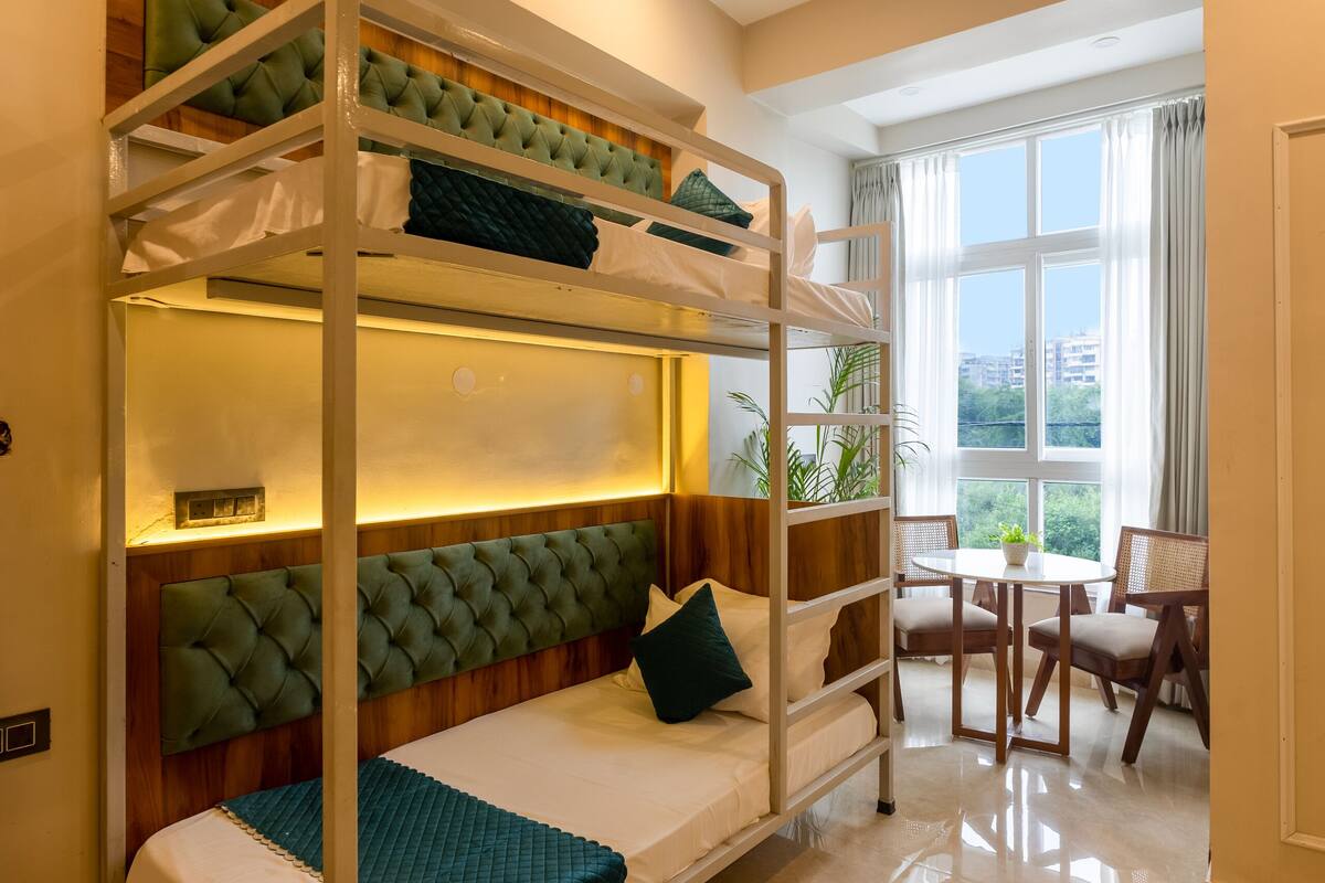 A bunk bed with a cushioned headboard, a small table with chairs, and a window with curtains.