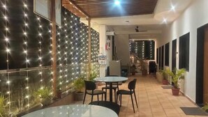 Un espace de restauration extérieur moderne, avec une table ronde et des chaises, entouré de plantes en pot et d’un mur décoré de guirlandes lumineuses.