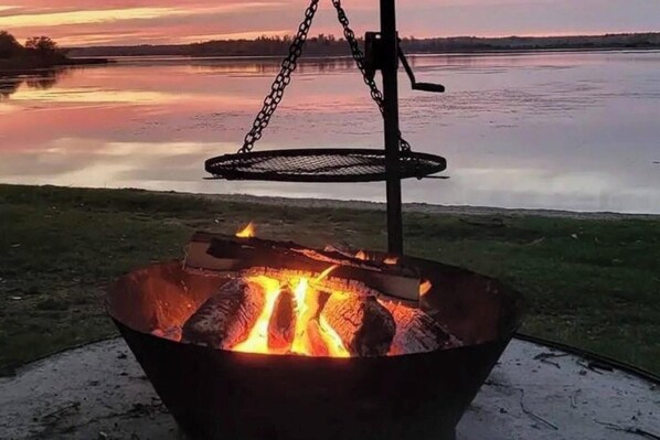 Un brasero de metal con una cadena y una rejilla, frente a un lago y un atardecer.