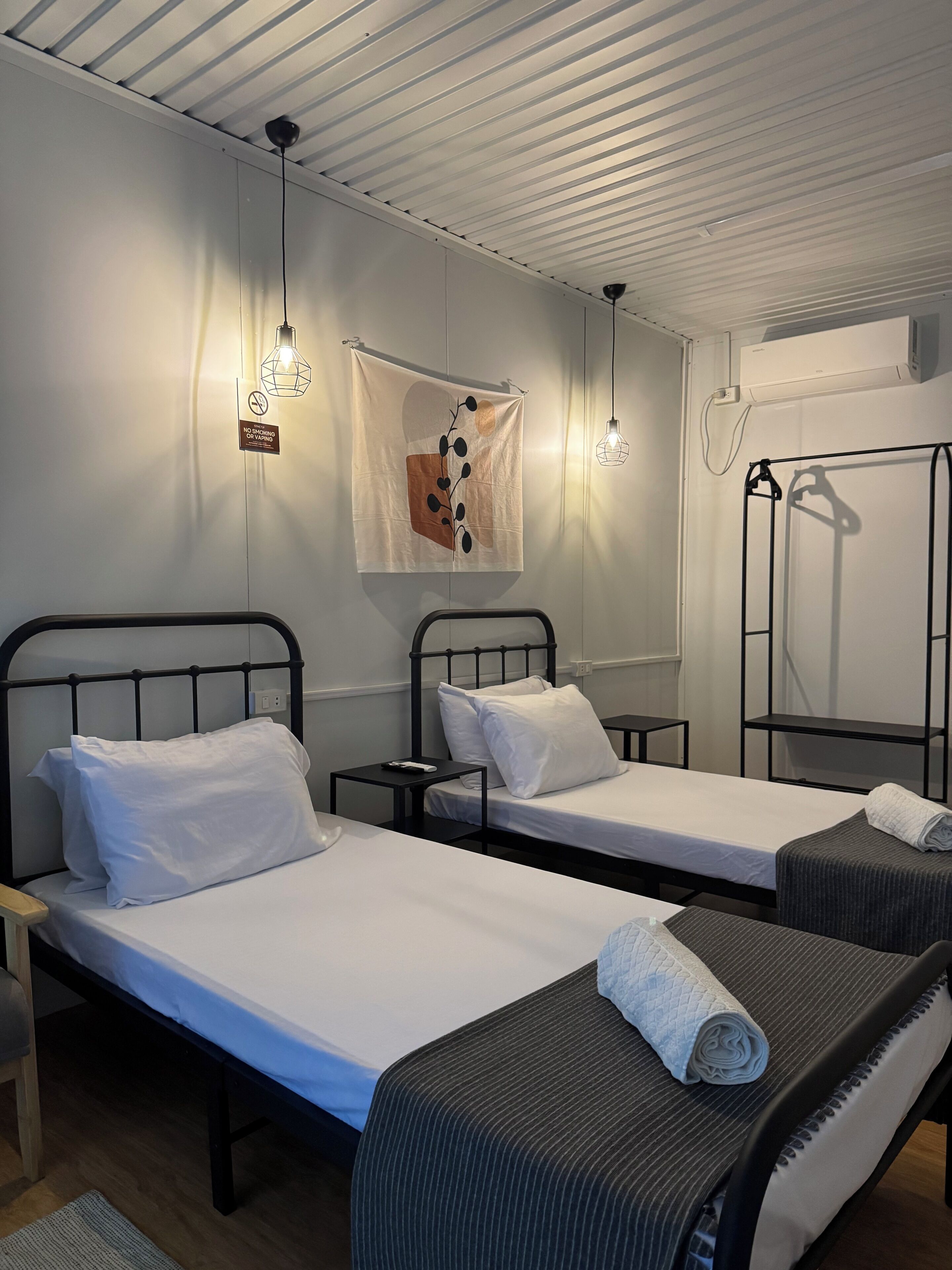 Ein Hotelzimmer mit zwei Einzelbetten, einem kleinen Tisch, einem Sessel, einer Pendelleuchte und einem Handtuchhalter.
