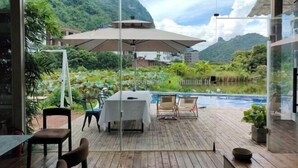 Una terraza de madera con una mesa y sillas, un paraguas grande y una piscina con montañas al fondo.