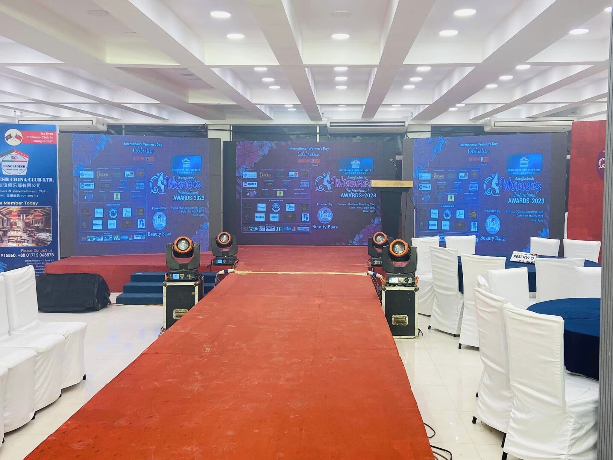 A conference room with a red carpet, white chairs, and large screens displaying the Bangladesh International Women's Day Awards 2023.
