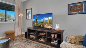 A living room with a TV, a cactus painting, and a framed artwork on the wall. - Modern 3BR House w/ Fast WiFi & Large Backyard (Tucson)
