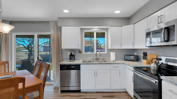 A modern kitchen with white cabinets, stainless steel appliances, and a dining area with wooden chairs and a table. - Modern 3BR House w/ Fast WiFi & Large Backyard (Tucson)