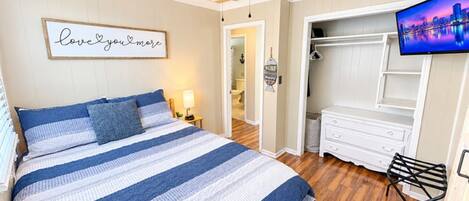 Una camera da letto con un letto, una ventola a soffitto, una TV, un armadio e un bagno visibile attraverso una porta aperta.
