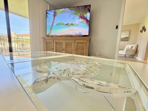 Ein modernes Wohnzimmer mit einem Glastisch, einem an der Wand montierten Flachbildfernseher und Blick auf einen Balkon mit Gartenmöbeln.