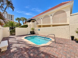Una piscina con patio de ladrillo, un edificio blanco con arcos y palmeras al fondo.