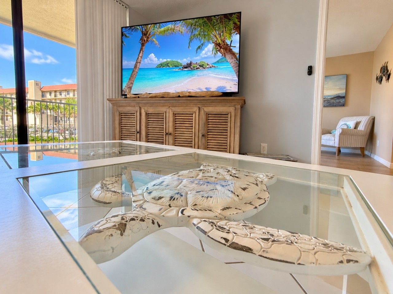 A modern living room with a glass-top coffee table, a flat-screen TV mounted on a wooden cabinet, and a view of a beach through large windows.