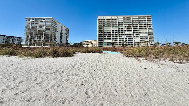 Un hotel frente a la playa con varios pisos y balcones, ubicado en una playa arenosa con poca vegetación.