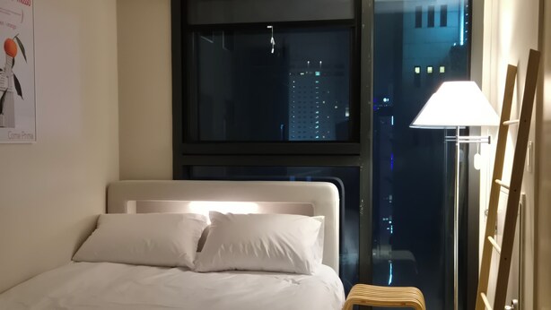 Hotellihuone, jossa on sänky, ikkuna, josta on näkymä kaupunkiin, lattiavalaisin ja juliste seinällä.