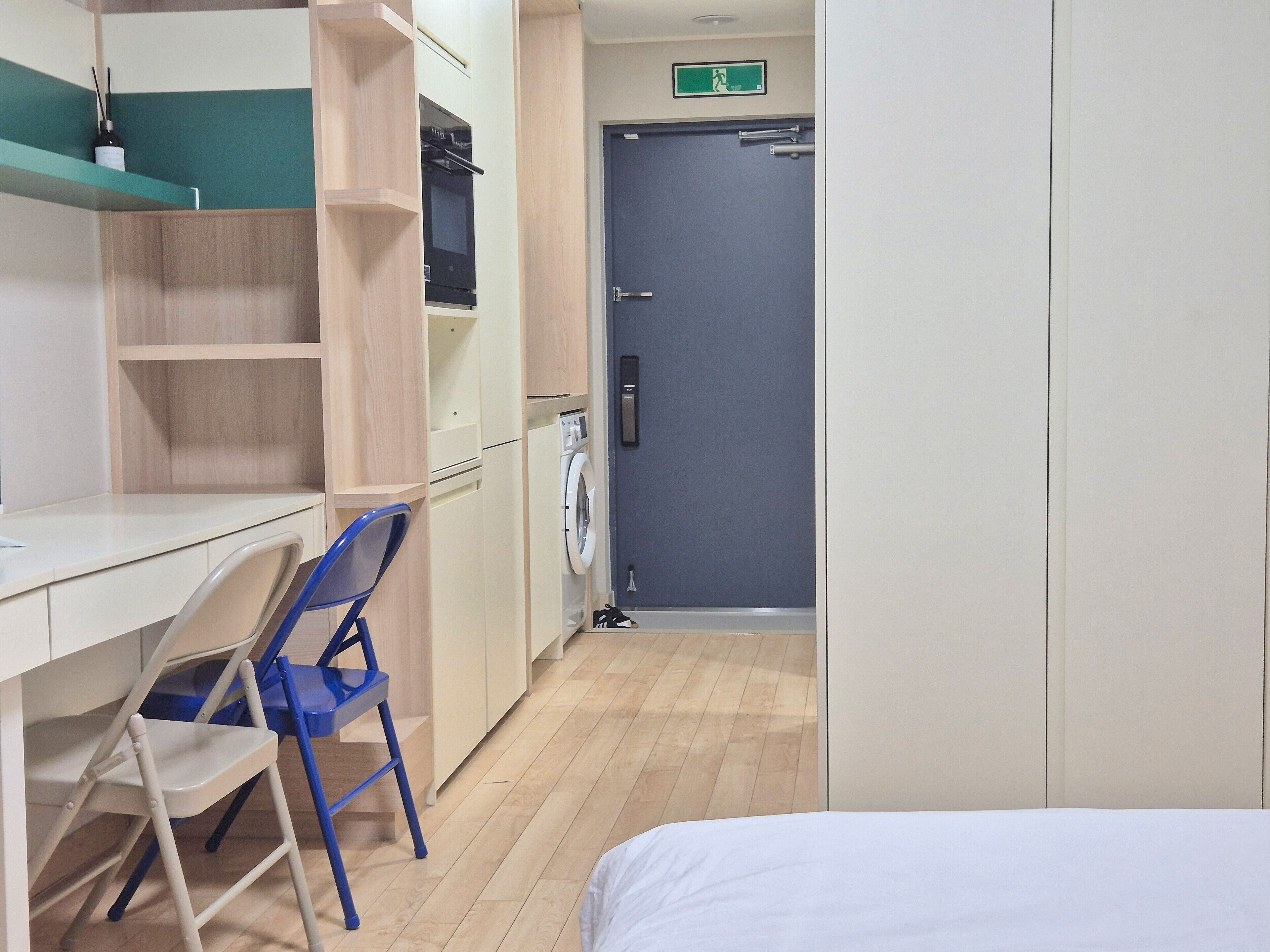 Una camera d'albergo compatta con un letto, una scrivania, una sedia, una lavatrice e una porta con il segnale di uscita di sicurezza.