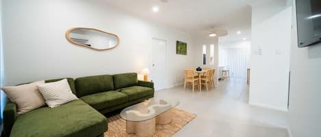 A modern living room with a green sofa, a glass coffee table, and a dining area with wooden chairs and a table.