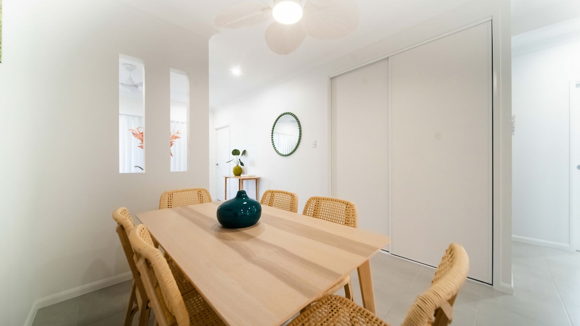 A dining area with a wooden table and wicker chairs, a green vase, and a wall-mounted mirror.