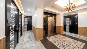 Een moderne hotel lobby met een centrale lift, een kamerbreed tapijt met patroon en zwart-wit deuren met kamer nummers.