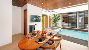 Un coin repas avec une table en bois dressée pour quatre personnes, un espace détente au bord de la piscine et une grande fenêtre donnant sur la piscine.
