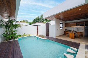 A modern outdoor pool area with a wooden deck, a dining area with a table and chairs, and a kitchen with white cabinets and a black refrigerator. - Villa Serene by Pertama Stay (Ungasan)