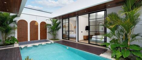 Un área de piscina moderna con cubierta de madera, un dormitorio con paredes de vidrio y plantas tropicales.