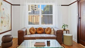 A living room with a leather sofa, a glass coffee table, and a view of buildings through a large window. - Belle Escapes - The Papilo (Holdfast Bay)