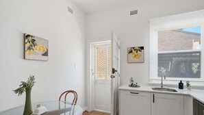 A compact kitchen with white cabinets, a glass dining table, and two paintings on the walls. - Belle Escapes - The Papilo (Holdfast Bay)