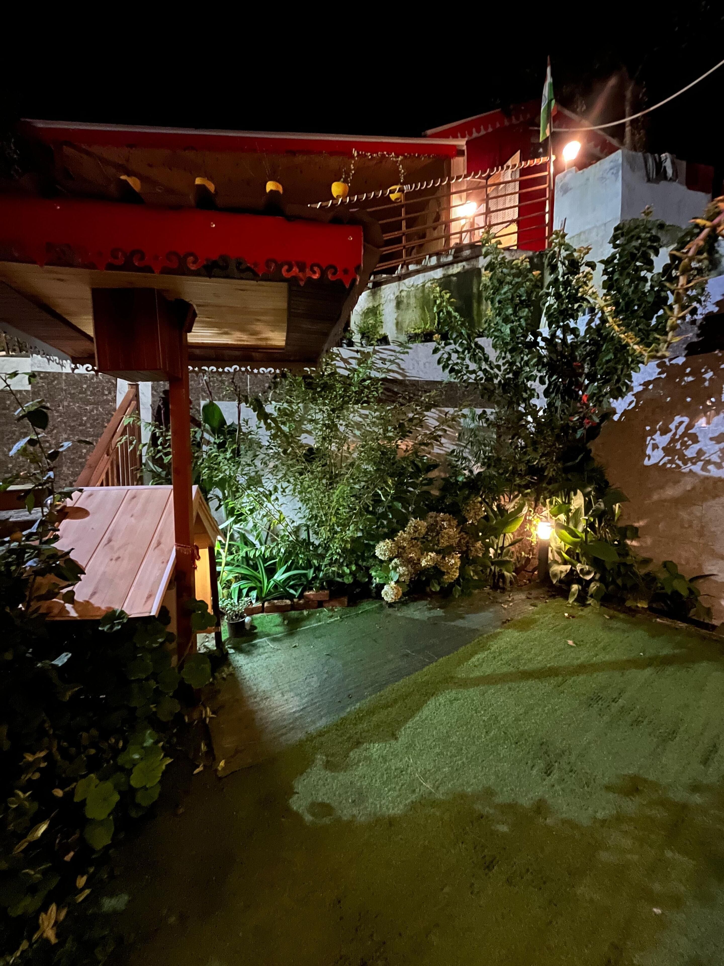 A nighttime view of a building with a red roof and a wooden deck. There are plants and a small garden area with a pathway leading to the deck.