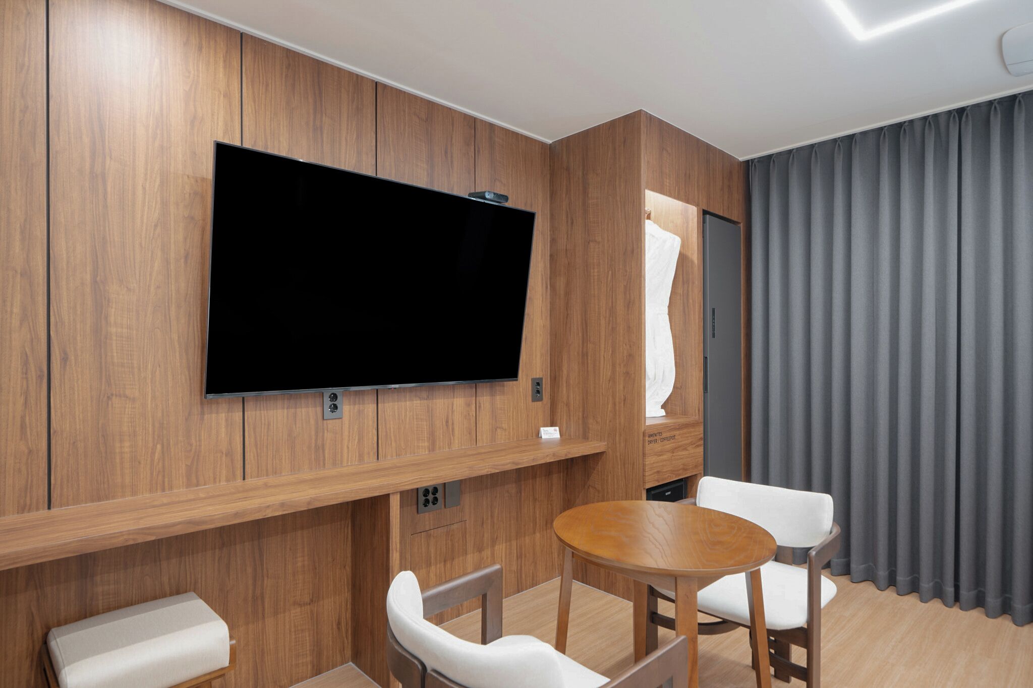 Une chambre moderne avec un téléviseur à écran plat fixé à un mur en bois, une table ronde en bois avec deux chaises blanches, un petit banc blanc et un rideau gris.