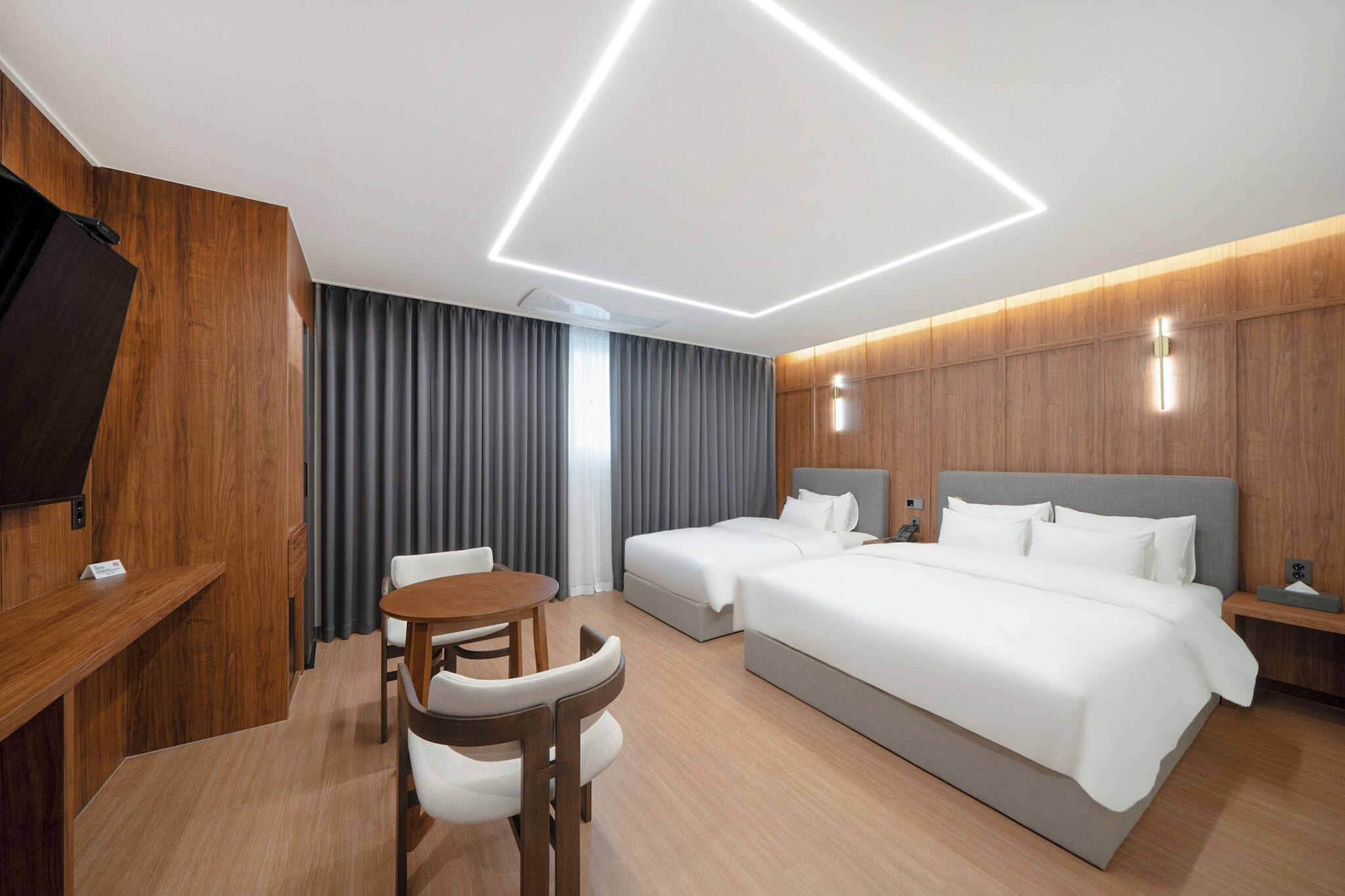 A modern hotel room with a wooden accent wall, a flat-screen TV, a desk, a round table, two beds, and a large window with curtains.