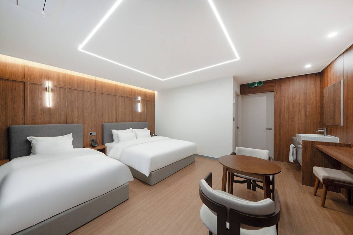 A modern hotel room with two beds, a dining area, and a bathroom.