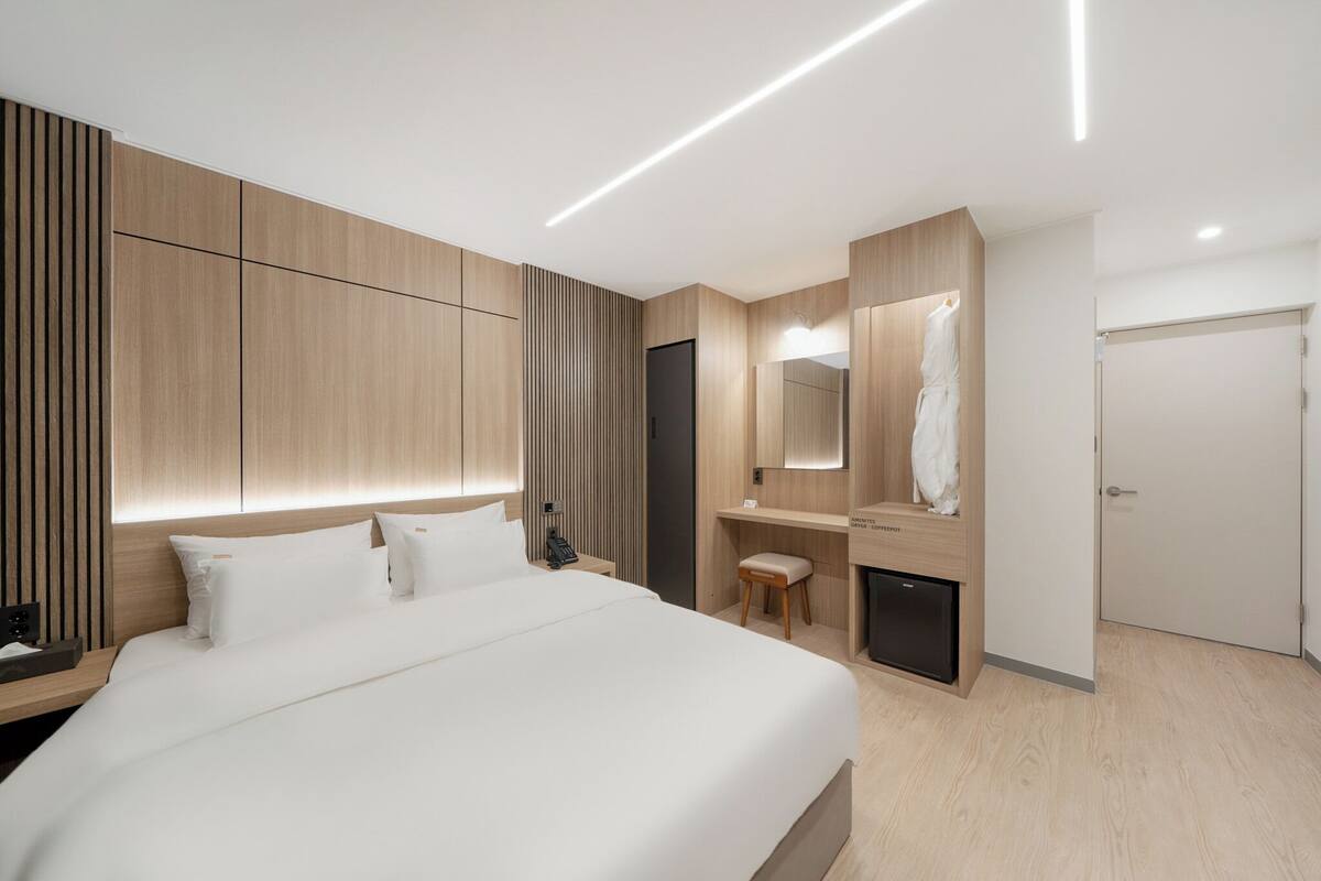 A modern hotel room with a large bed, built-in wardrobe, bedside table, and a small desk with a chair.