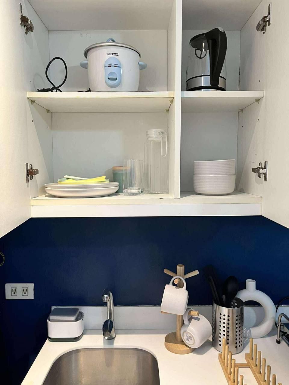 A kitchen with a sink, a coffee maker, a slow cooker, and a kettle on the shelves.