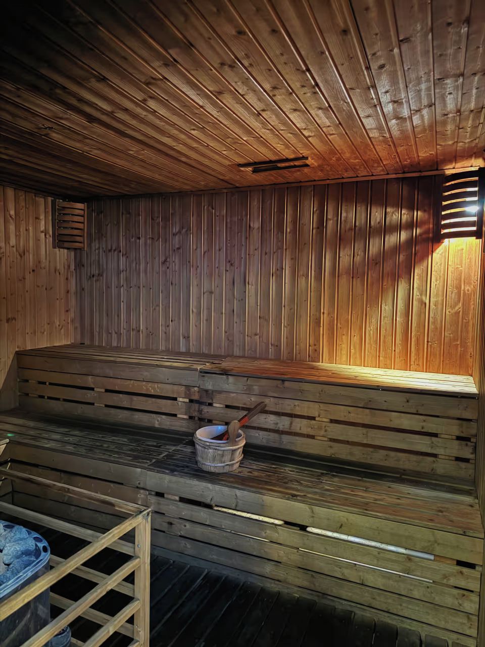 Egy szauna, melyben fából készült padok és egy vödör található, benne egy merőkanállal.