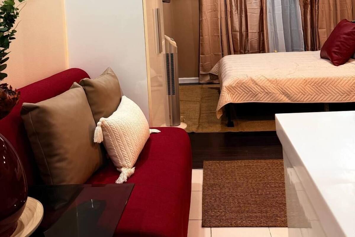 Een compacte hotelkamer met een rode bank, een klein tafeltje, een bed met een witte dekbedovertrek en een wandairconditioner.