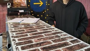 Une pièce avec une table dont le plateau est orné d’un motif en briques, une horloge, un tableau encadré et une porte décorée d’un cercle bleu et de points jaunes.