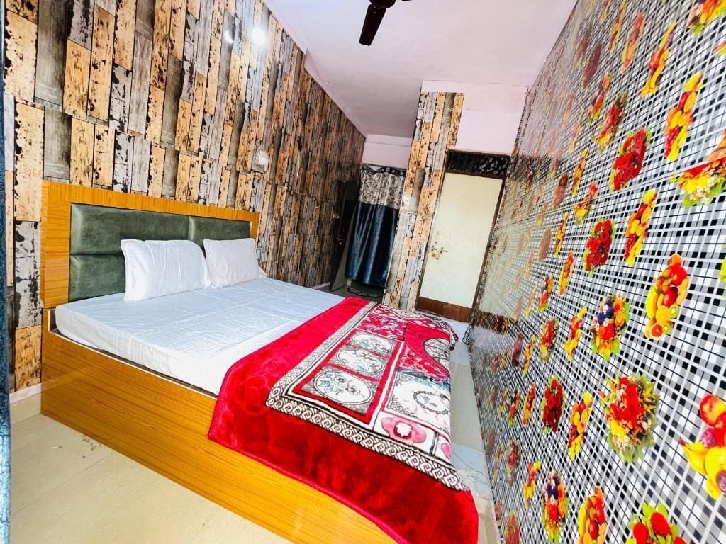Makuuhuone, jossa on puinen sängynpääty, valkoisilla tekstiileillä varustettu sänky, punakuvioinen heittopeitto ja kukka-asetelmalla koristeltu seinä.