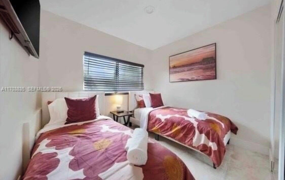 Ein Schlafzimmer mit zwei Betten, einem Fenster mit Jalousien, einem Bild an der Wand und einem kleinen Tisch.
