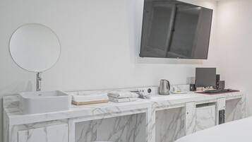 A modern bathroom with a marble vanity, a round mirror, a large flat-screen TV, and a desk with a computer setup.